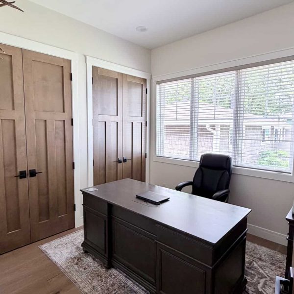 Modern home office with large window, black desk, and wooden closet doors.